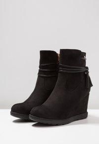 Stivaletti neri con tacco a zeppa realizzati in suede, dotati di una fascia in pelle avvolgente e un dettaglio a nappina, con una suola in gomma texturizzata per una buona aderenza.