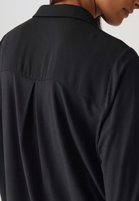 Camicia nera con una superficie liscia, dotata di un colletto a punta e un dettaglio sul retro plissettato. Il tessuto si drappeggia dolcemente.