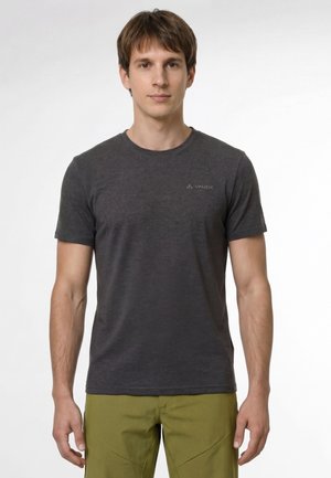 Giovane uomo che indossa una maglietta a maniche corte grigio scuro e pantaloni verde oliva, in piedi davanti a uno sfondo chiaro uniforme.