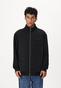 Black puffer jacket with a high collar, front zipper, and side pockets, featuring a quilted texture on the upper part and smooth sleeves.