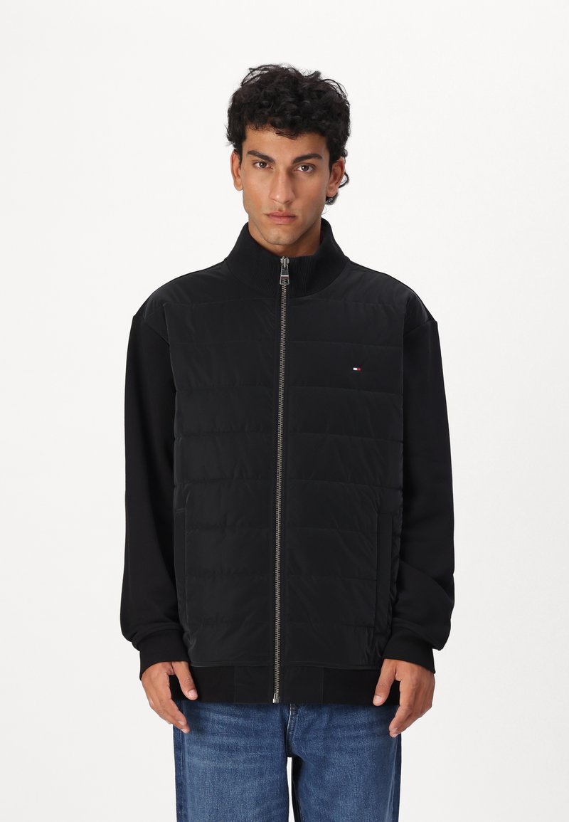 Black puffer jacket with a high collar, front zipper, and side pockets, featuring a quilted texture on the upper part and smooth sleeves.
