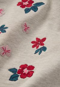 Lichtbeige stof met bloemenborduursel in rood, roze en groen. De bloemen variëren in grootte en detail, met weelderige bladeren die het ontwerp accentueren.