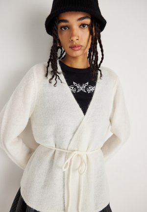 Cardigan en tricot doux et blanc, avec un design à col en V, des manches longues et une taille nouée. Porté sur une chemise noire avec un imprimé graphique.