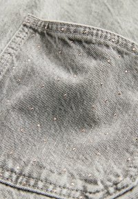 Gros plan d'une poche en tissu denim gris avec de petits clous argentés dispersés et des coutures doubles visibles.