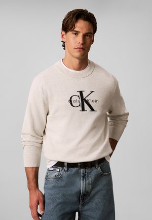 Άνδρας που φοράει ανοιχτό μπεζ πουλόβερ Calvin Klein και μπλε τζιν με μαύρη ζώνη, στέκεται μπροστά σε μονόχρωμο ανοιχτό γκρι φόντο.