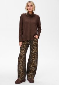 Kobieta stoi na białym tle w brązowym swetrze z golfem oraz szerokich spodniach w panterkę, nosząc ciemno-brązowe buty typu slip-on.