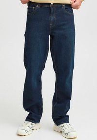 Jeans de mezclilla azul oscuro con corte recto, cinco bolsillos y costuras naranjas contrastantes. Llevados con zapatillas deportivas blancas.