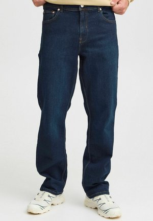 Mørkeblå denimjeans med rett ben, fem lommer og kontrasterende oransjefargede sømmer. Brukt med hvite treningssko.