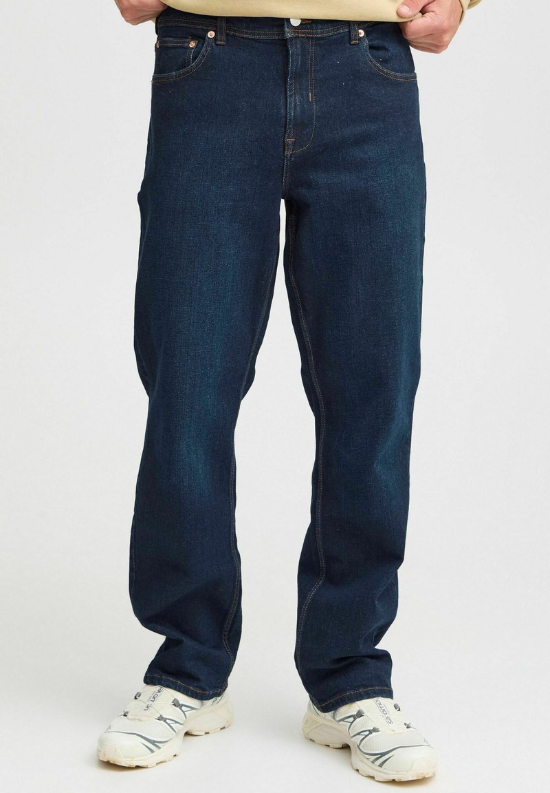 Jeans de mezclilla azul oscuro con corte recto, cinco bolsillos y costuras naranjas contrastantes. Llevados con zapatillas deportivas blancas.