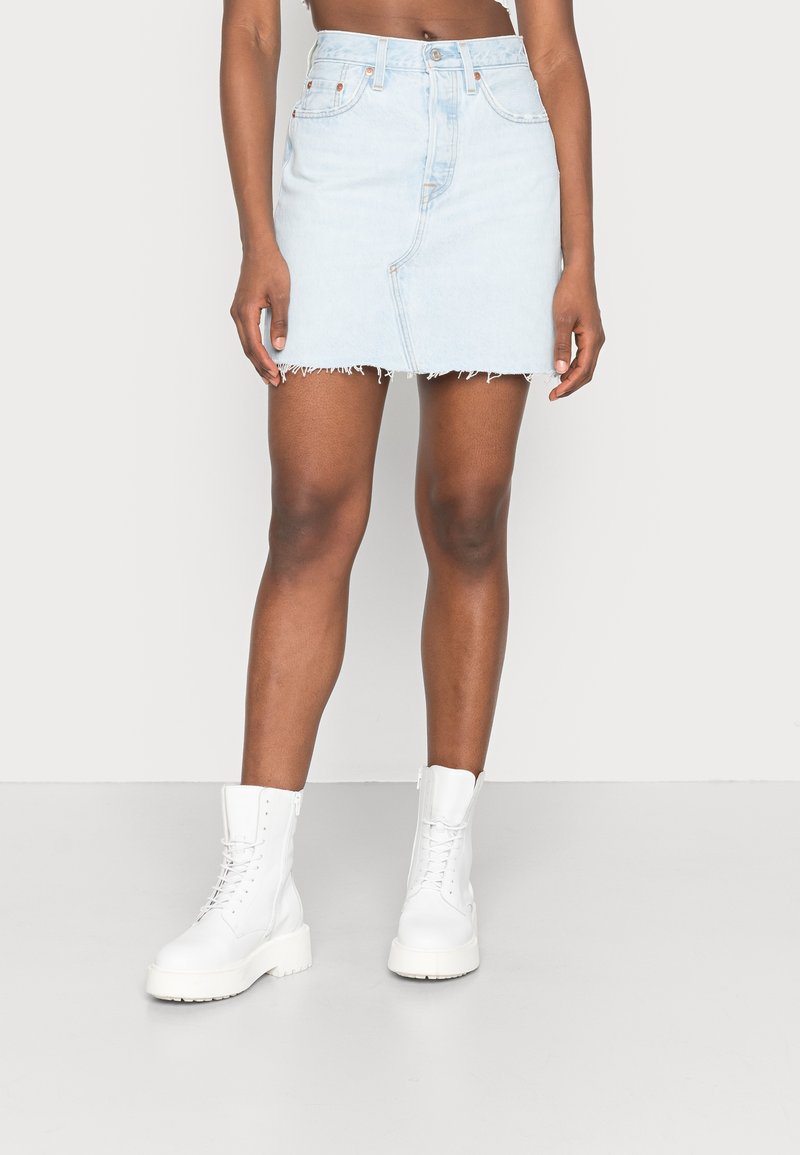 Kvinna i ljusblå denim-minikjol med rå fåll och vita snörade kängor som står på ett vitt golv.