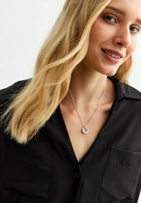 Zilveren halsketting met een ronde hanger aan een dunne ketting, gedragen over een zwarte knoopshirt. De hanger heeft twee in elkaar grijpende ringen.