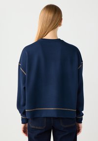 Granatowy, oversize'owy sweter z długimi rękawami, z ribbowymi mankietami i poziomym, złotym akcentem wyszytym na plecach. Gładka tekstura, luźny krój.