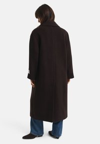 Lange, donkerbruin wollen jas met een brede kraag, rechte snit, lange mouwen en een opvallende naaddetail aan de achterkant. Gepaarde met blauwe jeans.