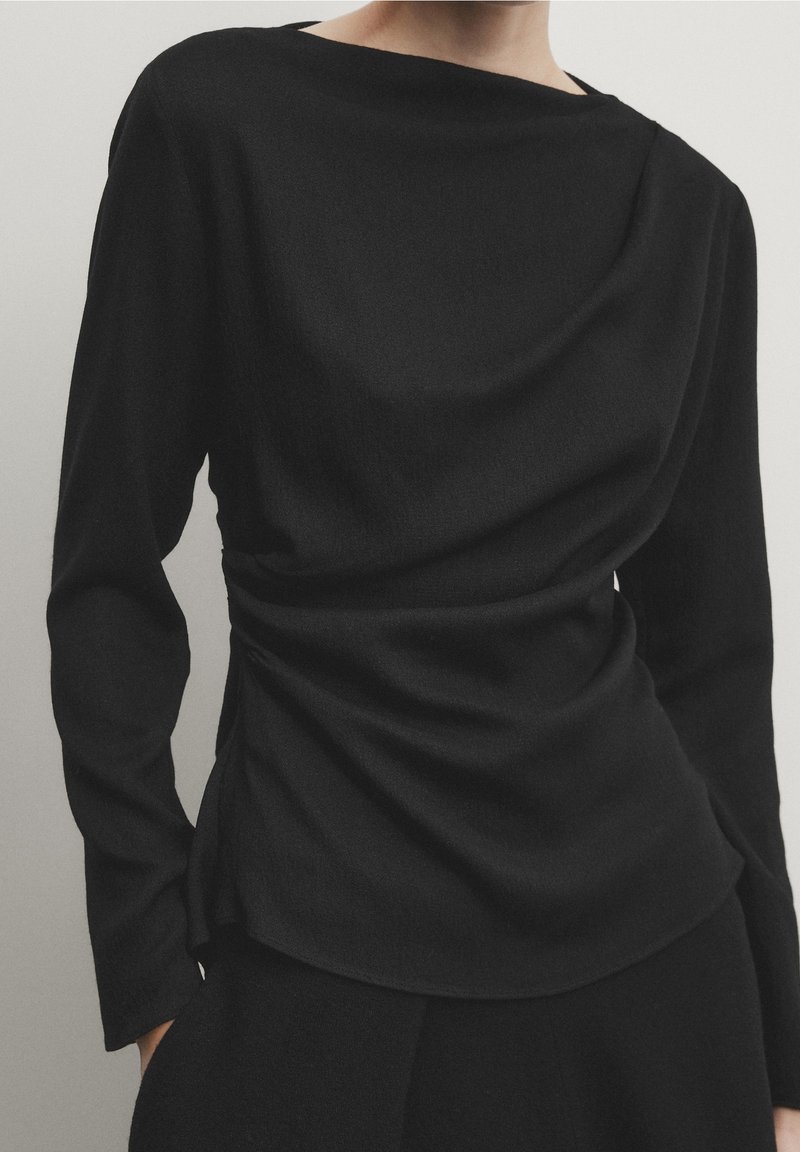 Black long-sleeve top with a draped design and wide neckline. Soft fabric features subtle texture and gathers at the waist for a fitted look.