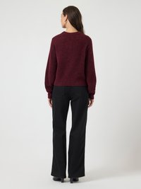 Žena stojaca chrbtom v tmavočervenom svetríku s dlhými rukávmi, čiernych nohaviciach a čiernych lodičkách na jednoduchom svetlom pozadí.