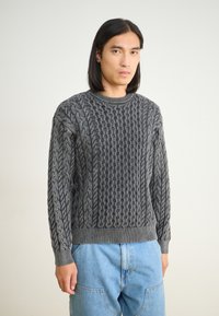 Szürke, cable-knit pulóver texturált mintával, kerek nyakkivágással és bordázott mandzsettákkal, világoskék farmernadrággal párosítva.