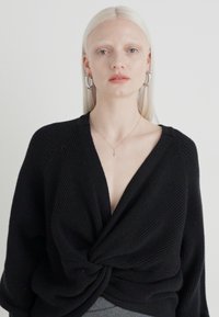 Pulover negru tricotat cu decolteu adânc în V și detaliu frontal răsucit. Textură côtă, croială lejeră și mâneci lungi. Design simplu, fără modele.