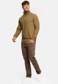 Sudadera de felpa de color verde oliva con cuello tipo zip hasta el cuarto, combinada con jeans slim-fit marrones y zapatillas blancas, de pie contra un fondo claro.