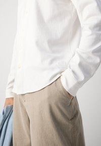 Witte lange mouwen button-up shirt met een ronde zoom en een logo-tag. Gecombineerd met beige broeken, die een gladde textuur en een ontspannen pasvorm uitstralen.