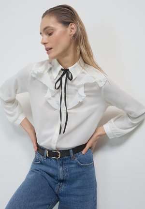 White, semi-sheer blouse with ruffled accents and a black tie at the neck, paired with high-waisted blue jeans and a black belt.