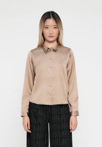 Camicia button-up in satin beige con colletto a coste e maniche lunghe, caratterizzata da una vestibilità dritta e una leggera lucentezza. Indossata con pantaloni neri testurizzati.