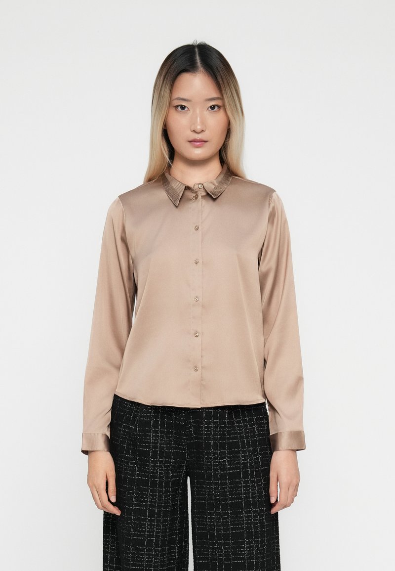 Camicia button-up in satin beige con colletto a coste e maniche lunghe, caratterizzata da una vestibilità dritta e una leggera lucentezza. Indossata con pantaloni neri testurizzati.