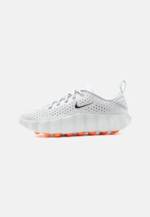 Biały sneaker Nike z perforowaną cholewką, srebrnym logo Swoosh, sznurowanym zapięciem, grubą podeszwą i pomarańczowymi teksturowanymi poduszkami trakcyjnymi pod spodem.