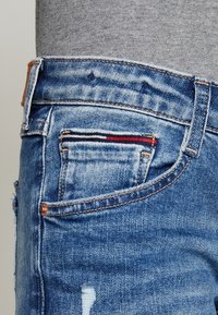 Jeans de mezclilla con un lavado azul claro, que presentan áreas desgastadas y un patrón en rojo, blanco y azul en el bolsillo trasero.