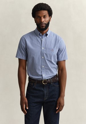 Mann mit Bart, der ein kurzärmliges blau kariertes Hemd mit Knöpfen, dunkle Jeans und geflochtenen Ledergürtel trägt und vor einfachem Hintergrund steht.