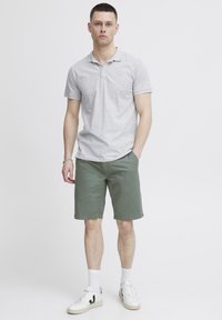 Camisa polo gris, pantalones cortos verdes y zapatillas blancas. Mangas cortas, tapeta de botones en la camisa, corte casual y diseño sencillo.