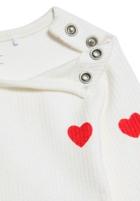 Weiße gerippte Stoff mit roten Herzmustern an den Ärmeln und silbernen Druckknöpfen am Ausschnitt. Weicher Textur, lässiges Design.