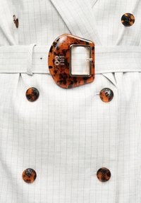 Άσπρο παλτό με ρίγες, με κουμπιά από χελώνα και ταιριαστό αγκράφα ζώνης με μεταλλικό ορθογώνιο πλαίσιο και αρχικά της μάρκας.