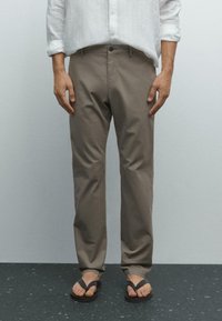 Man die staat en beige broeken draagt, een wit overhemd met opgerolde mouwen en zwarte slippers op een donker gevlekte vloer.