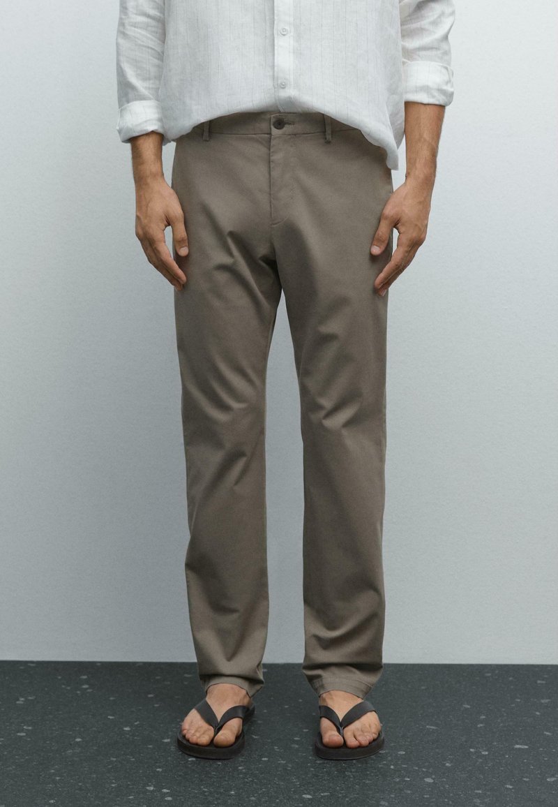 Man die staat en beige broeken draagt, een wit overhemd met opgerolde mouwen en zwarte slippers op een donker gevlekte vloer.