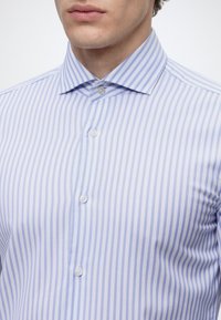 Homme portant une chemise boutonnée avec des rayures verticales bleu clair et blanches et un col pointu.