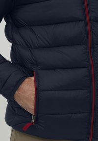 Mørkeblå pufferjakke med røde glidelåsdetaljer og quiltet tekstur. Har en sidelomme og en tett passform ved kragen og mansjettene.