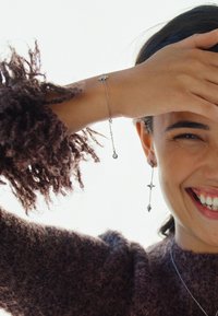 Gioielli in argento con un bracciale a catena con una gemma centrale e lunghi orecchini pendenti con dettagli a forma di stella, indossati con un maglione marrone testurizzato.