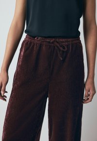 Bordo spalvoti, tekstūriniai poilsio kelnės, pagamintos iš minkštos medžiagos, turi elastingą juosmenį ir surišimo detalę. Kirpimas – laisvas.