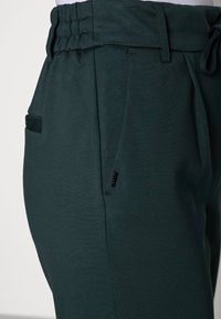 Dark green jogger pants with a soft texture, featuring a drawstring waistband, side pockets, and a small brand tag on the side.