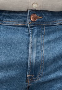 Sinised teksapüksid, millel on esiküljel vaskne nööp ja nähtav oranž õmblus, kantud beeži särgiga, mis on sisse topitud.