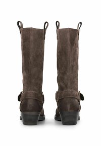 Cizme călduroase din suede maro, până la genunchi, cu toc tip platformă, accentuate de o curea cu cataramă. Au două bucle de prindere în partea de sus pentru a fi ușor de încălțat.