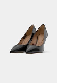 Une paire d'escarpins en cuir verni noir à bout pointu avec des semelles intérieures beige et des détails discrets de logo de designer sur un fond blanc.