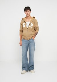 Svetlohnedá mikina na zips s bielym logom "GAP", v kombinácii so svetlomodrými rozšírenými džínsami a bielymi teniskami, na jednolatom pozadí.