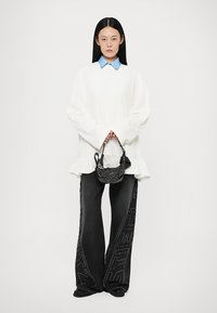 White long-sleeve blouse with frilled hem, layered over black wide-leg pants featuring a textured logo. Small woven black handbag with accents.