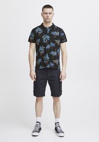 Svart polo t-skjorte med fargerik palmeprint, sammen med marineblå cargo-shortser. Modellen har på seg svarte høyhælte sneakers. En enkel, uformell antrekk.