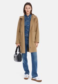 Beige Wollmischungsmantel mit Kapuze, Vordertaschen und Kordelzug. Darunter eine blaue Jeansjacke und blaue Jeans mit weißen Sneakers.