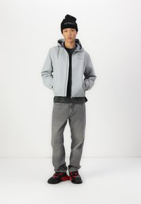 Lichtgrijze zip-up jack met capuchon, zwart t-shirt eronder, grijze jeans en zwarte schoenen met rode accenten. Zwarte beanie op het hoofd.