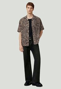 Jovem em pé, vestindo uma camisa de leopardo de mangas curtas sobre uma blusa preta, calças pretas e sapatos pretos, em fundo simples.