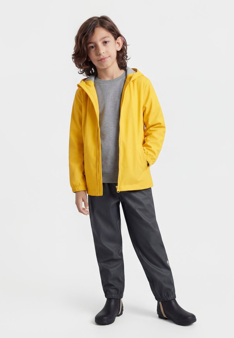 Bambino con capelli castano chiaro fino alle spalle che indossa una giacca impermeabile gialla, felpa grigia, pantaloni impermeabili neri e stivali neri, in piedi su sfondo bianco.