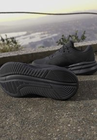 Paire de chaussures de sport noires sur un pavement texturé avec une chaussure montrant le motif de la semelle et un arrière-plan scénique flou.